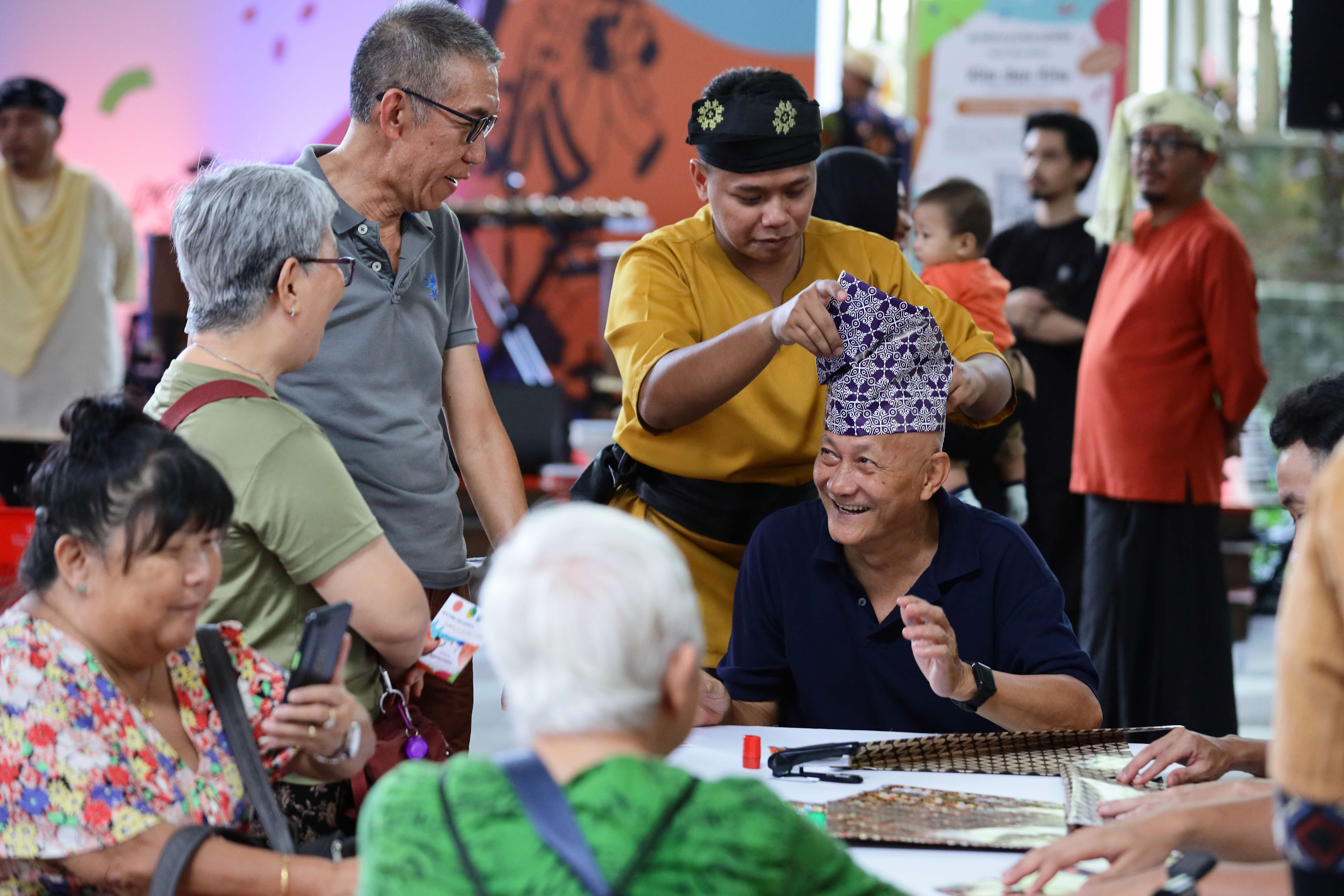 A man fits a batik hat on another man's head. Other people watch and participate.